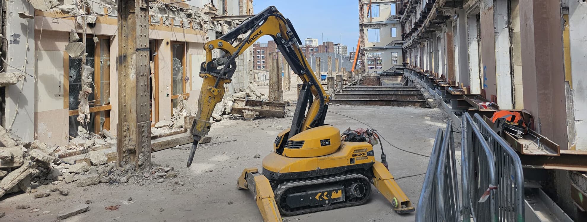 Yellow Brokk robotic demolition machine working inside a partially-demolished heritage building, the listed facade retained around it while the internal concrete structure comes out cleanly — top-down deconstruction under a facade-retention scheme