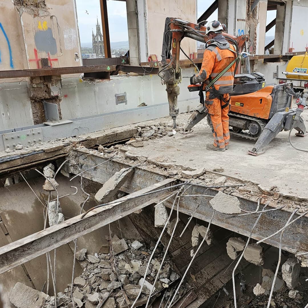 GNAT UK operator with a Brokk pecker breaking out the deck of a heritage building, listed cathedral spire visible through the cleared facade — top-down robotic deconstruction under conservation control