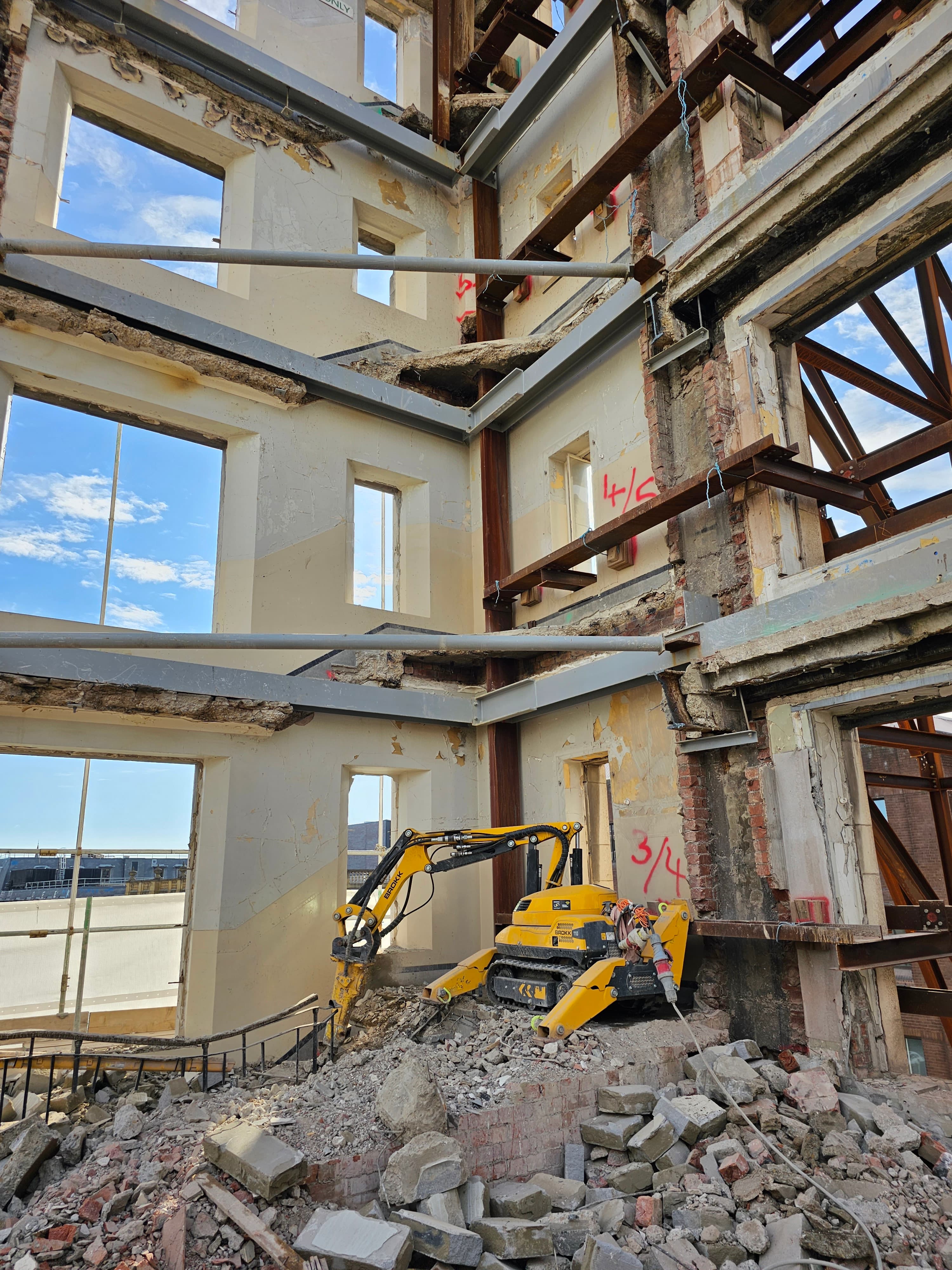 Yellow Brokk demolition robot working through the upper-floor structure of a Georgian-shell building, original sash-window facade preserved while the post-war concrete intervention comes out