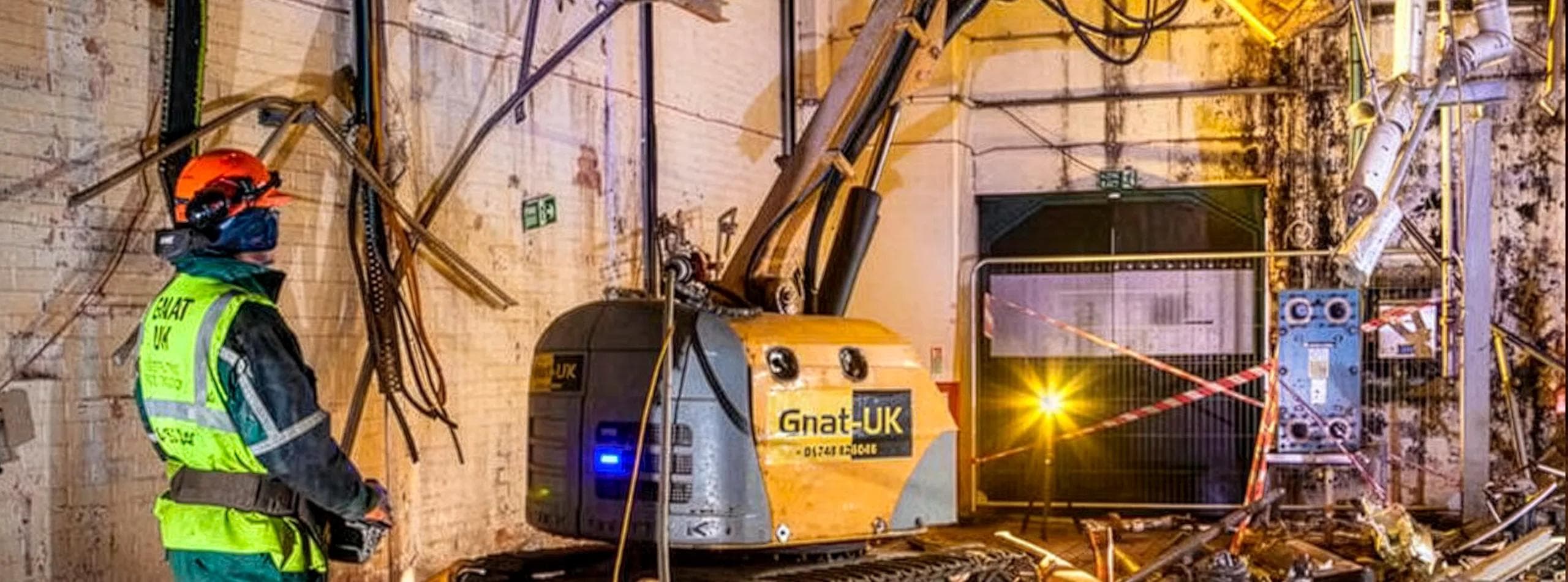 Brokk 330 robotic demolition machine deployed inside a factory roof void — GnatUK-branded plant in the foreground with Atlas Copco steel shears attachment, hi-vis operator observing remotely, twisted pipework being severed in the dimly-lit industrial space