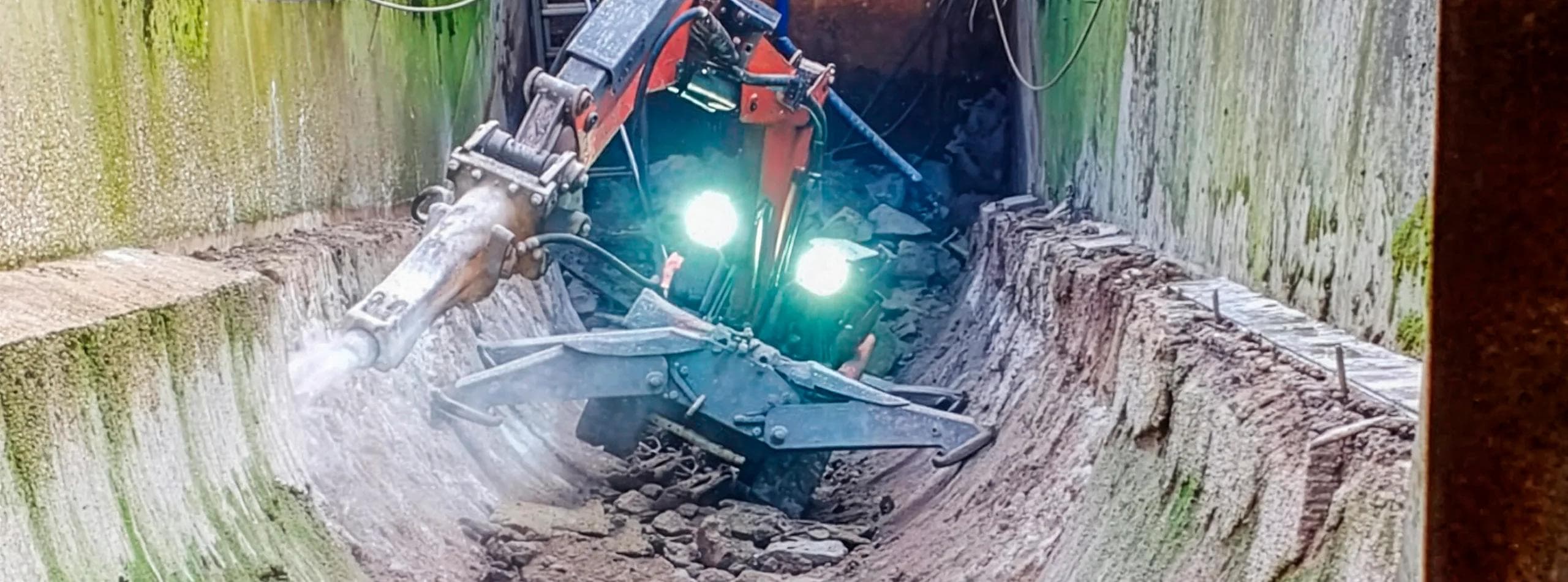Husqvarna DXR140 robot inside a 40-degree Archimedes screw trough excavation — green algae-stained walls of the operational asset, twin work-lights illuminating the working face, concrete screed being selectively removed