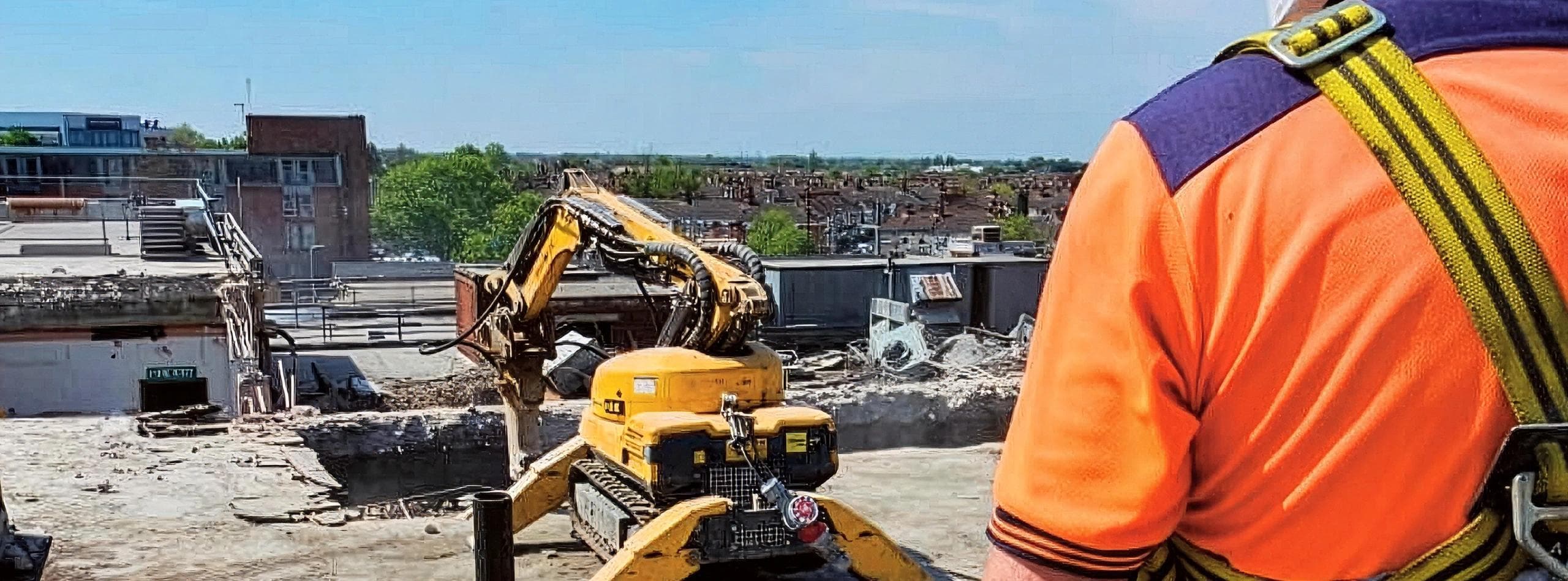 Top-down robotic demolition in progress on the Leeds tower — Brokk excavator perched on a partially-deconstructed rooftop floor, GNAT UK operator in orange hi-vis and full harness watching from the working edge with the Leeds residential rooftops stretching out behind